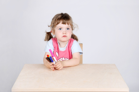 Little sad girl sits at wooden table and holds with felt-tip pens on grey background.の写真素材