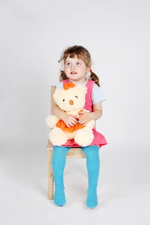 Little cute girl sits at wooden chair with toy bear and looks away on grey background.の写真素材