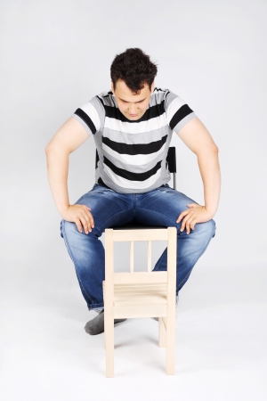 Brunet handsome man sits on chair and looks small wooden chair on grey background.の写真素材