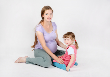 Pregnant smiling woman sits on floor with little daughter on grey background.の写真素材