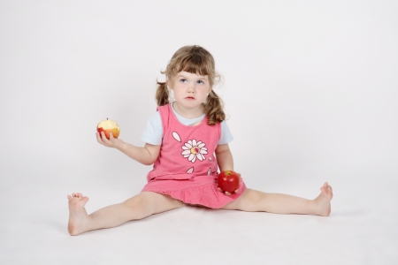 Little beautiful girl eats apple and holds second apple on floor in hand on white background.の写真素材
