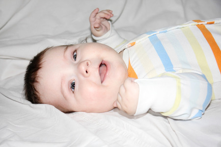 Little beautiful baby in striped jumper lies and smiles on white sheet on bed.の写真素材