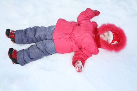 Happy little girl in warm clothes lies in snow in winter day.の写真素材