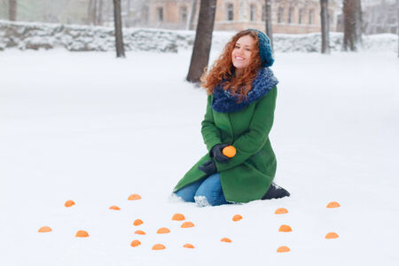 Beautiful girl sits in snow among mandarins at winter day in parkの写真素材