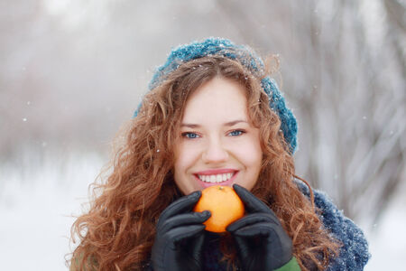Beautiful girl in blue beret holds mandarin and smiles outdoor at winter day in parkの写真素材