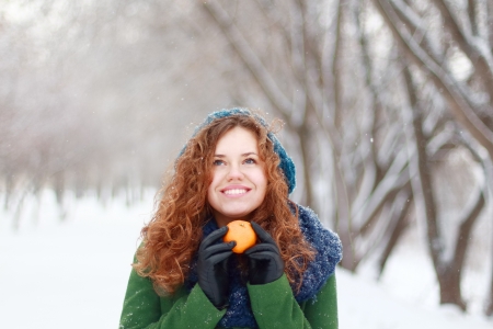 Beautiful girl in blue beret holds mandarin and looks up at winter day in parkの写真素材