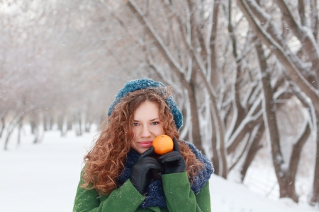 Beautiful girl holds mandarin and looks at camera at winter day in parkの写真素材