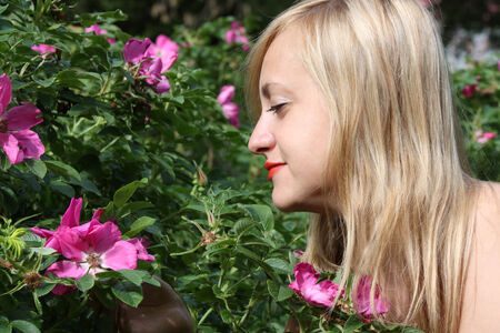 Beautiful blonde girl looks at pink flowers on bush in park at summer dayの写真素材
