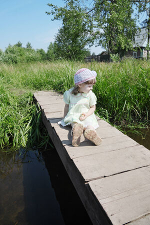 Pretty little girl sits on small bridge in village at summer sunny dayの写真素材