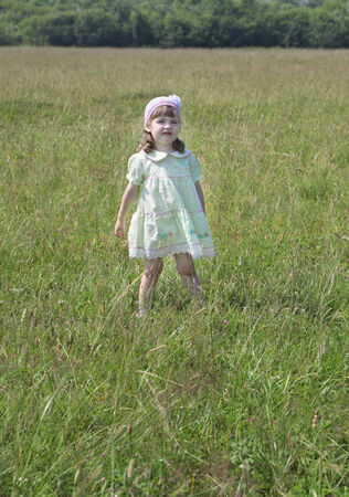 Cute little girl in dress stands at green meadow at summer sunny dayの写真素材