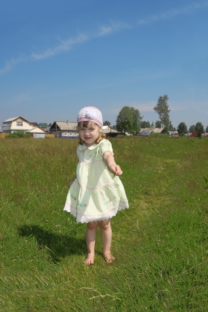 Little barefoot girl plays shows blade of grass at green meadow near village at summer sunny dayの写真素材