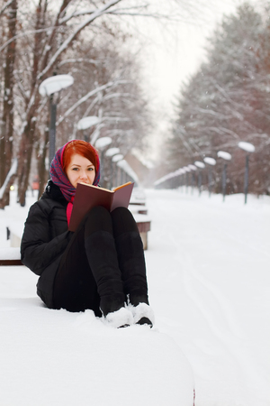 Beautiful girl in black sits on bench with book outdoor at winter day in parkの写真素材