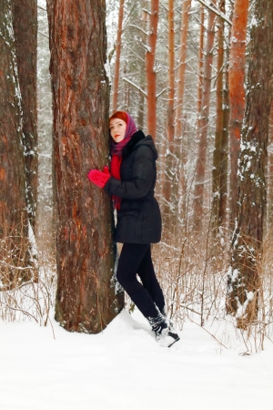 Beautiful girl stands next to large tree and looks up outdoor at winter day in forestの写真素材