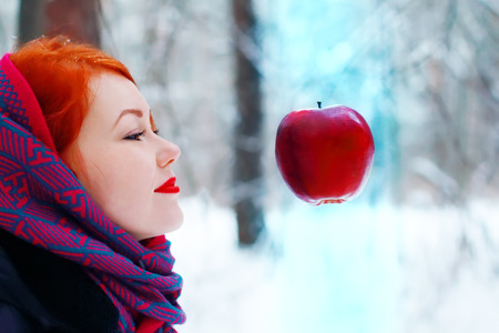 Smiling girl looks at hanging in air big red apple outdoor at winter day in forest