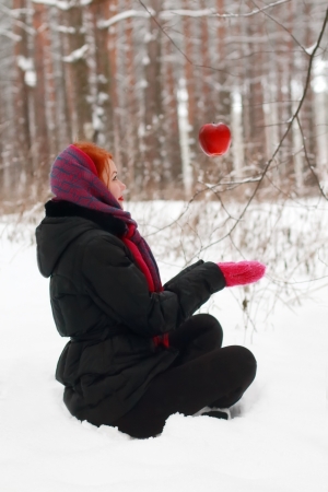 Smiling girl sits on snow and looks at hanging in air big red apple outdoor at winter day in forestの写真素材