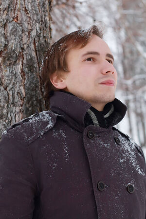 Young man with snowflakes in hair stands near tree and looks up at winter snowy dayの写真素材