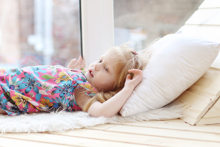 Pretty little blonde girl lies on white pillow near window in room and looks upの写真素材