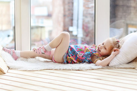 Pretty little blonde girl laughs and lies on white pillow and fur near window in roomの写真素材