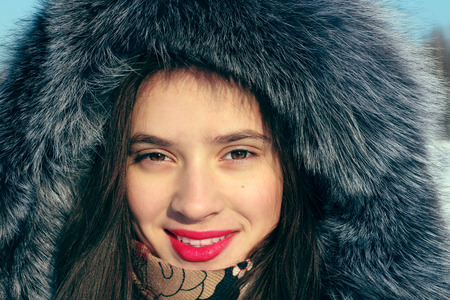 Beautiful girl in fur coat with hood smiles and looks at camera in sunny winter day