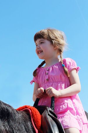 Little beautiful girl in pink dress sits in saddle on brown horseの写真素材