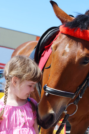 Little beautiful girl in pink dress stands near brown horse at sunny dayの写真素材