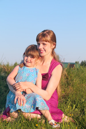 Smiling mom with her daughter sitting on grass in summer sunny dayの写真素材