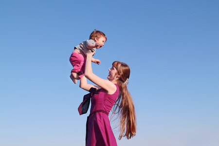 Beautiful smiling young woman in pink dress holding her son on sky backgroundの写真素材
