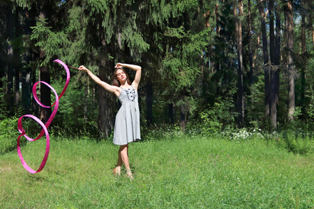 Beautiful girl in dress is engaged in rhythmic gymnastics with ribbon on grassの写真素材