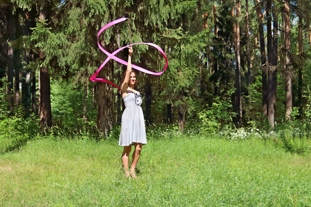 Beautiful girl in dress, is engaged in rhythmic gymnastics with ribbon on grass in forestの写真素材