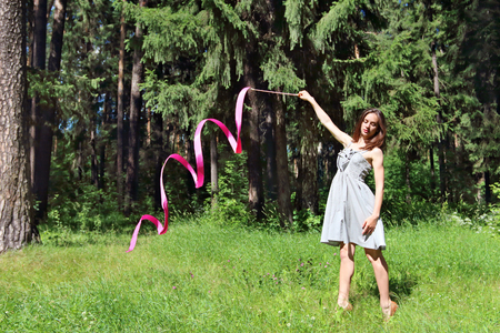 Beautiful girl in dress, is engaged in rhythmic gymnastics with ribbon on grass in woods on summer dayの写真素材