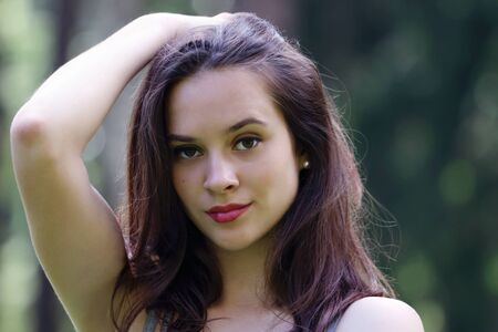 Close-up face of beautiful girl straightens hair with her hand with view of forest in background with shallow depth of fieldの写真素材