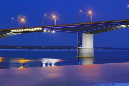 Part of bridge with lanterns and reflection in water of river with ice and snow at winter nightの写真素材