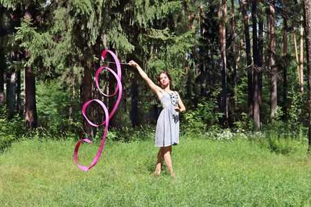 Beautiful girl in dress is engaged in rhythmic gymnastics with ribbon on grass on sunny summer dayの写真素材