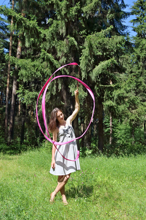 Beautiful girl in dress, is engaged in rhythmic gymnastics with ribbon in woods on summer dayの写真素材