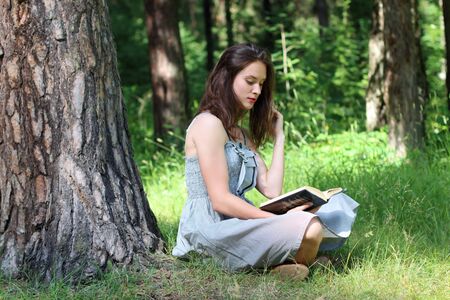 Beautiful girl in dress sitting under tree on grass and reading bookの写真素材