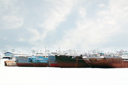 Old and rusty big freight ships on frozen river at winter cloudy dayの写真素材