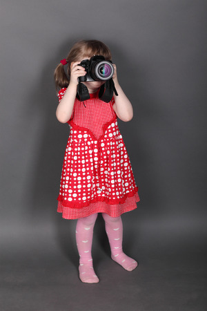 Funny little girl in red dress shoots by modern camera in grey studioの写真素材