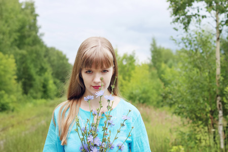 Beautiful woman in blue holds chicory flowers and smiles among trees at overcast dayの写真素材