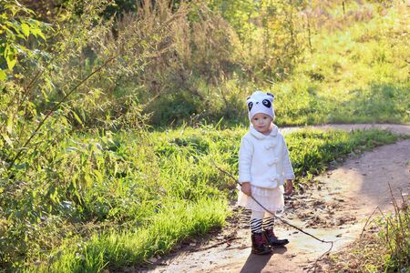 Pretty little girl in white stands with stick on path in park in sunlightの写真素材