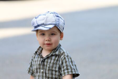 Handsome little boy in cap stands on asphalt and looks away at summer dayの写真素材