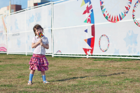 Happy little girl in skirt licks lollipop on grass outdoor in townの写真素材