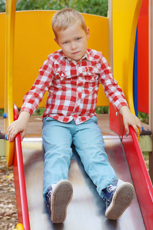 Handsome little boy in shirt sits on slide on playground in autumn dayの写真素材