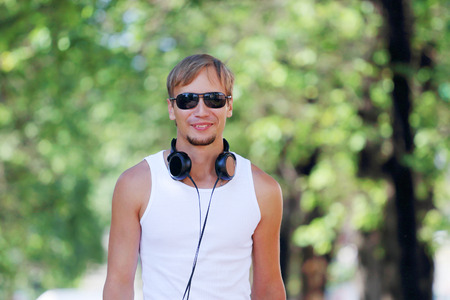 Smiling man in sunglasses and headphones on alley with green trees at summer dayの写真素材