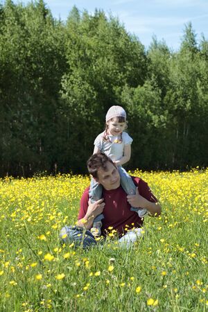 Happy father with his daughter on shoulders among yellow flowers at meadow at summer dayの写真素材