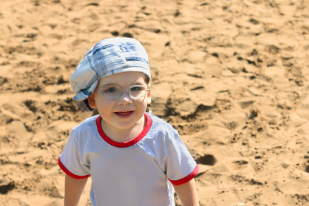 Little handsome boy in cap and glasses smiles on sand at summer dayの写真素材
