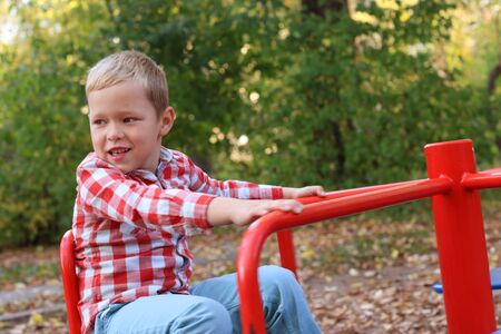 Handsome little boy in shirt plays on small carousel on playground in autumn dayの写真素材
