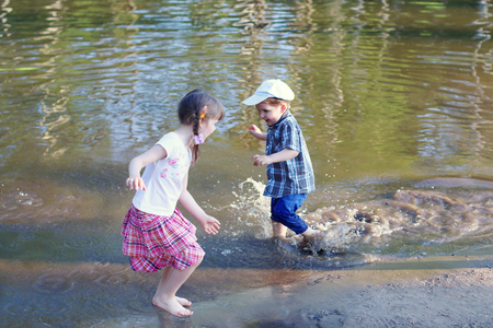 Little barefoot girl with boy laugh and run in water of pond at summer dayの写真素材