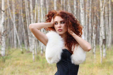 Young woman in corset with fur and curly hair looks away in autumn forestの写真素材