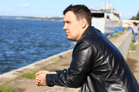Man in leather jacket stands near river with ship, looks away and dreamsの写真素材