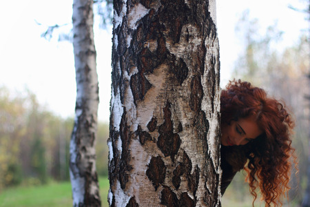 Pretty smiling woman looks out of big birch in autumn forest, shallow dofの写真素材